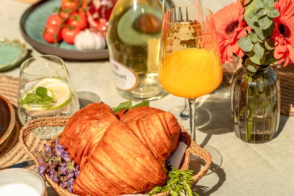 A table with croissants, a glass of orange juice, a bottle of wine, and a vase with flowers.