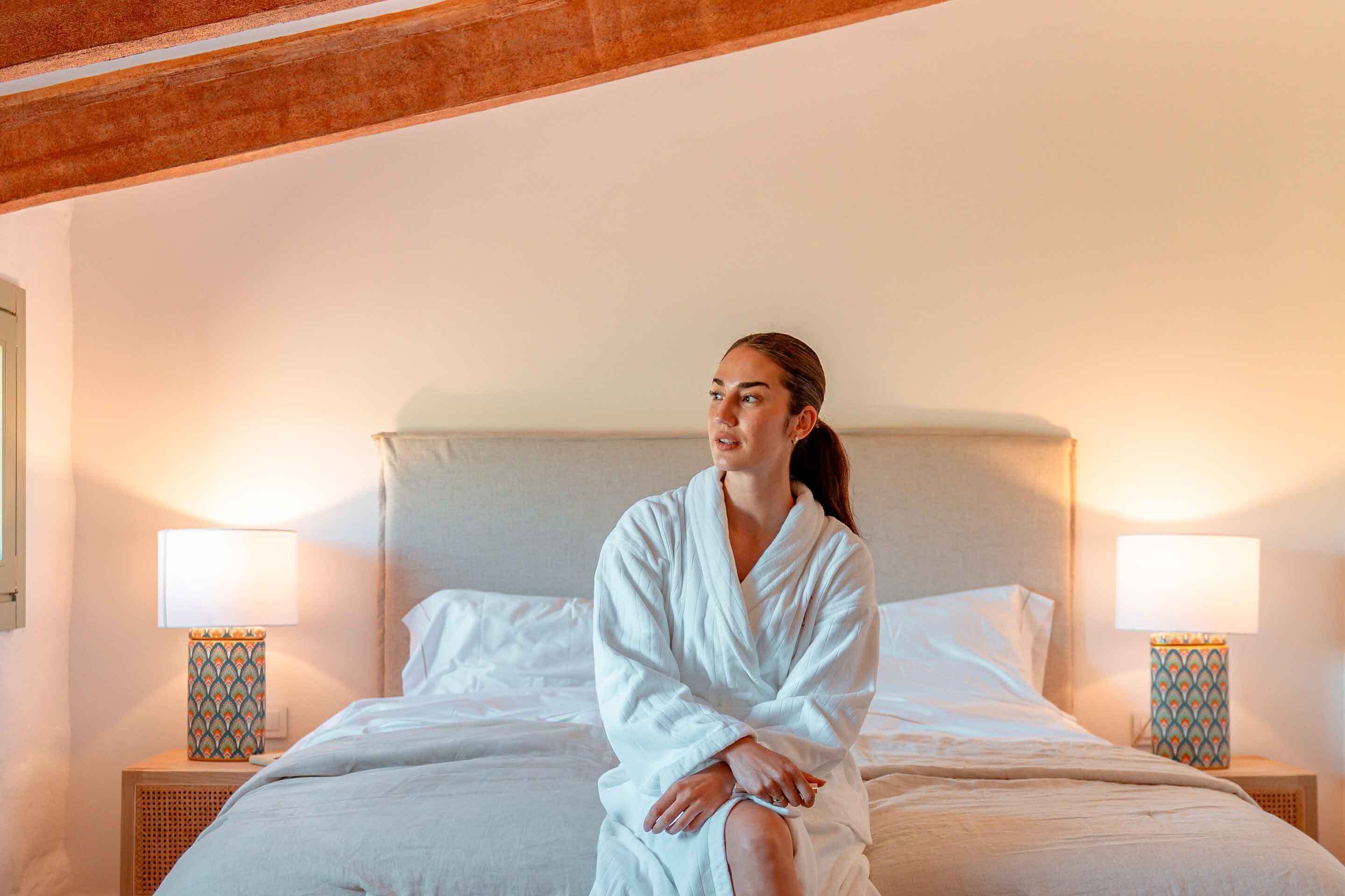 Une personne est assise sur un lit dans une pièce dont le plafond est en bois, qui est équipée de deux lampes de chevet et dont un des murs est uni.