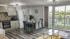 Eine moderne Küche mit weißen Schränken, einer Mikrowelle und einem Herd. Ein Essbereich mit Tisch für vier Personen, Stühlen und einer Pflanze. Eine Uhr hängt an der Wand, und das Zimmer hat große Fenster mit Blick ins Freie.