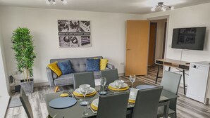 Ein modernes Wohnzimmer mit Essbereich, ein graues Sofa mit blauen und gelben Kissen, eine Topfpflanze und ein an der Wand befestigtes Fernsehgerät.