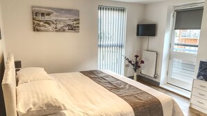 Ein Schlafzimmer mit Bett, Fernseher, Fenster mit Jalousien, Heizkörper und einem Bild an der Wand.