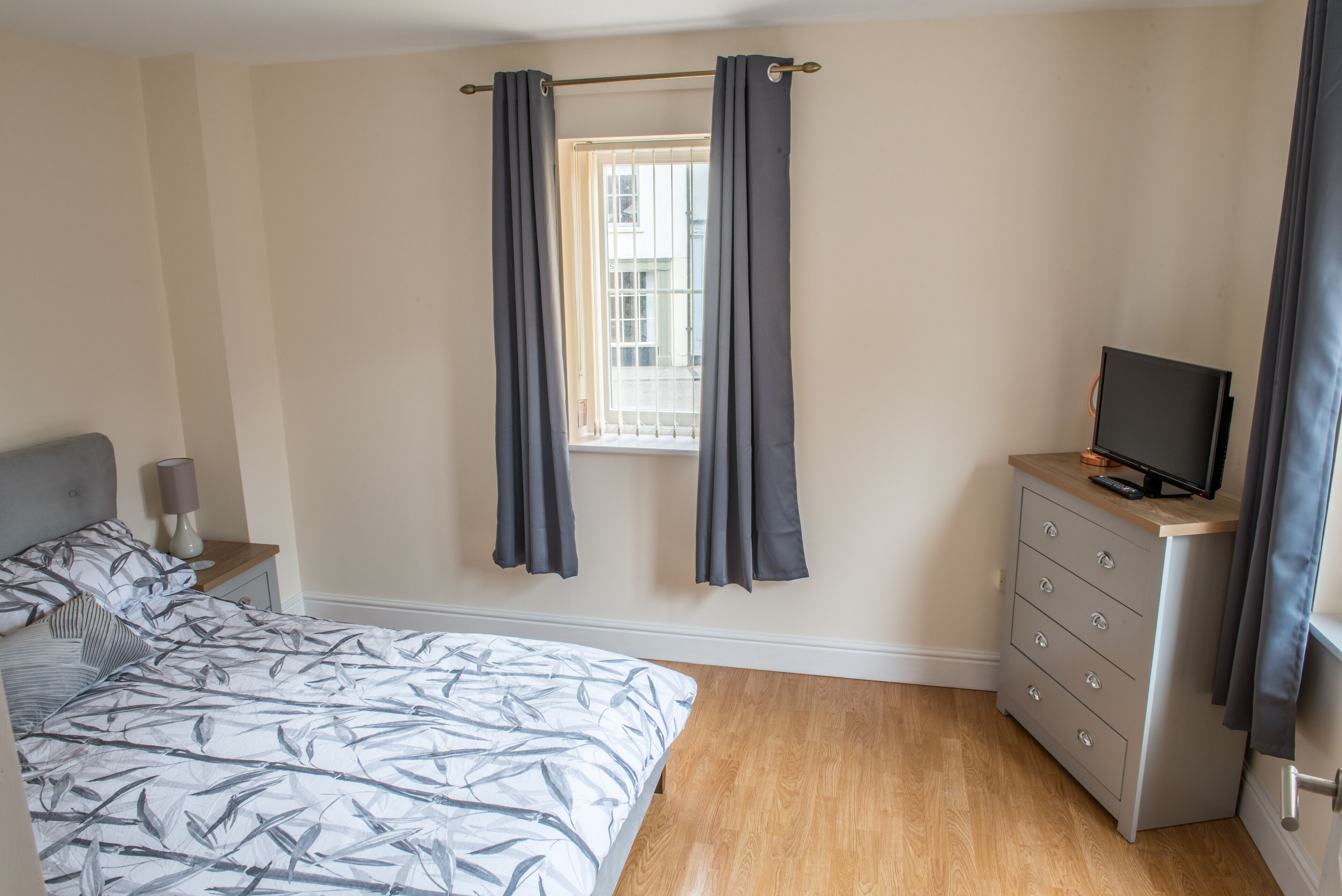Een slaapkamer met een bed, een kaptafel, een televisie en een raam met gordijnen.