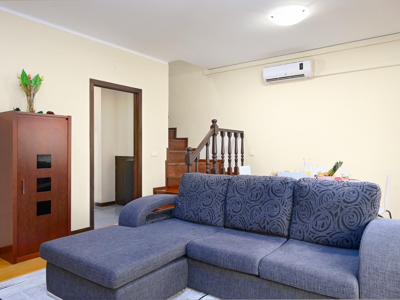 En stue med en grå hjørnesofa, et træskab, en trappe og en aircondition-enhed på væggen.