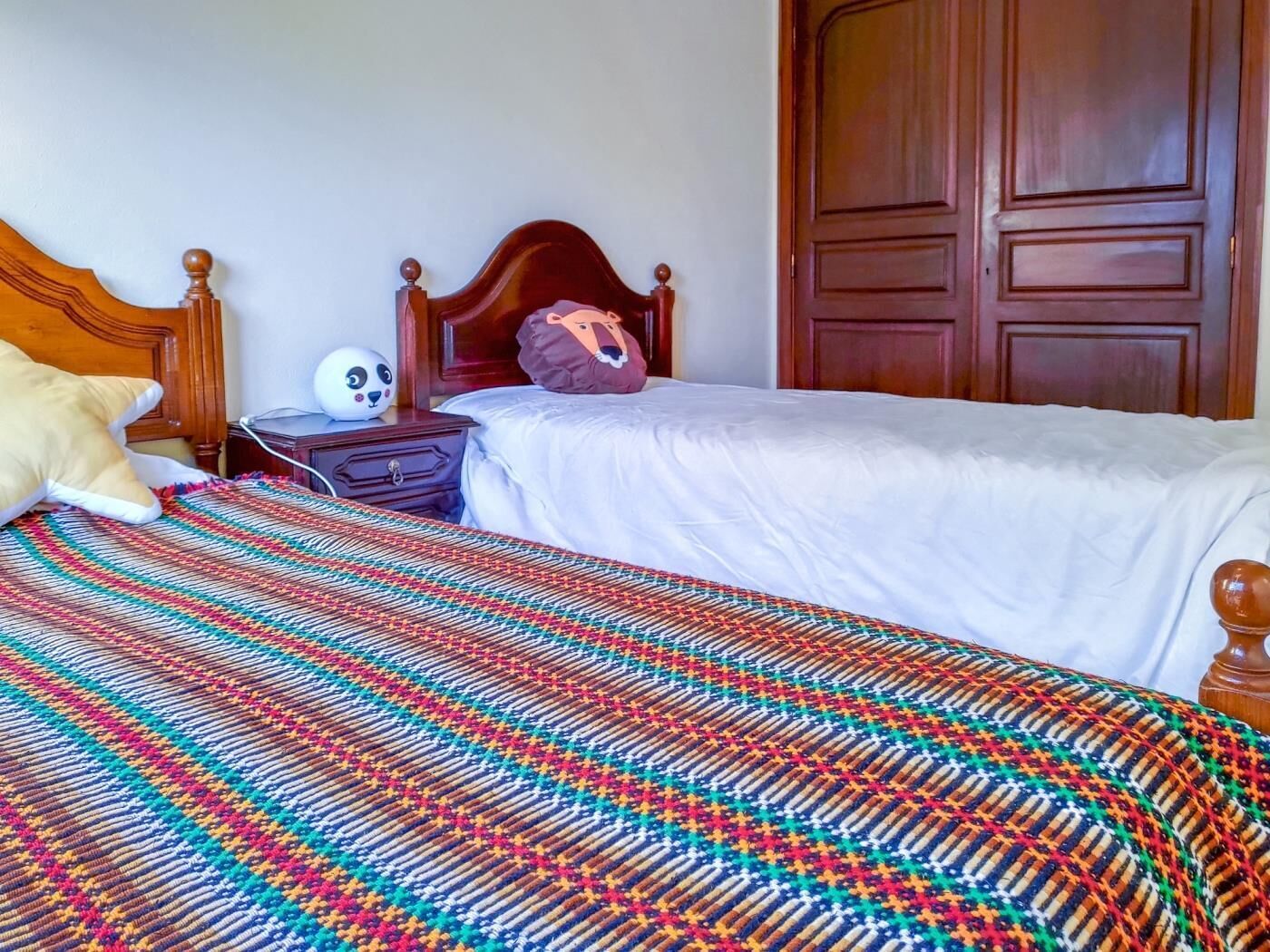 Habitación de hotel con una cama de madera, una manta tejida de colores, una mesita de noche con lámpara y un armario.