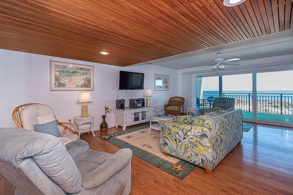 A spacious living room with a wooden ceiling, a large TV, a comfortable sofa, and a view of the beach through large windows.