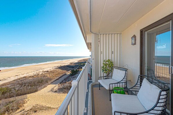 A balcony with a view of the beach, a chair, a small table, and a potted plant.