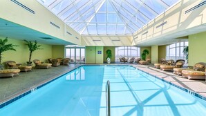 Indendørs swimmingpool med liggestole og plantekrukker, omgivet af store vinduer.