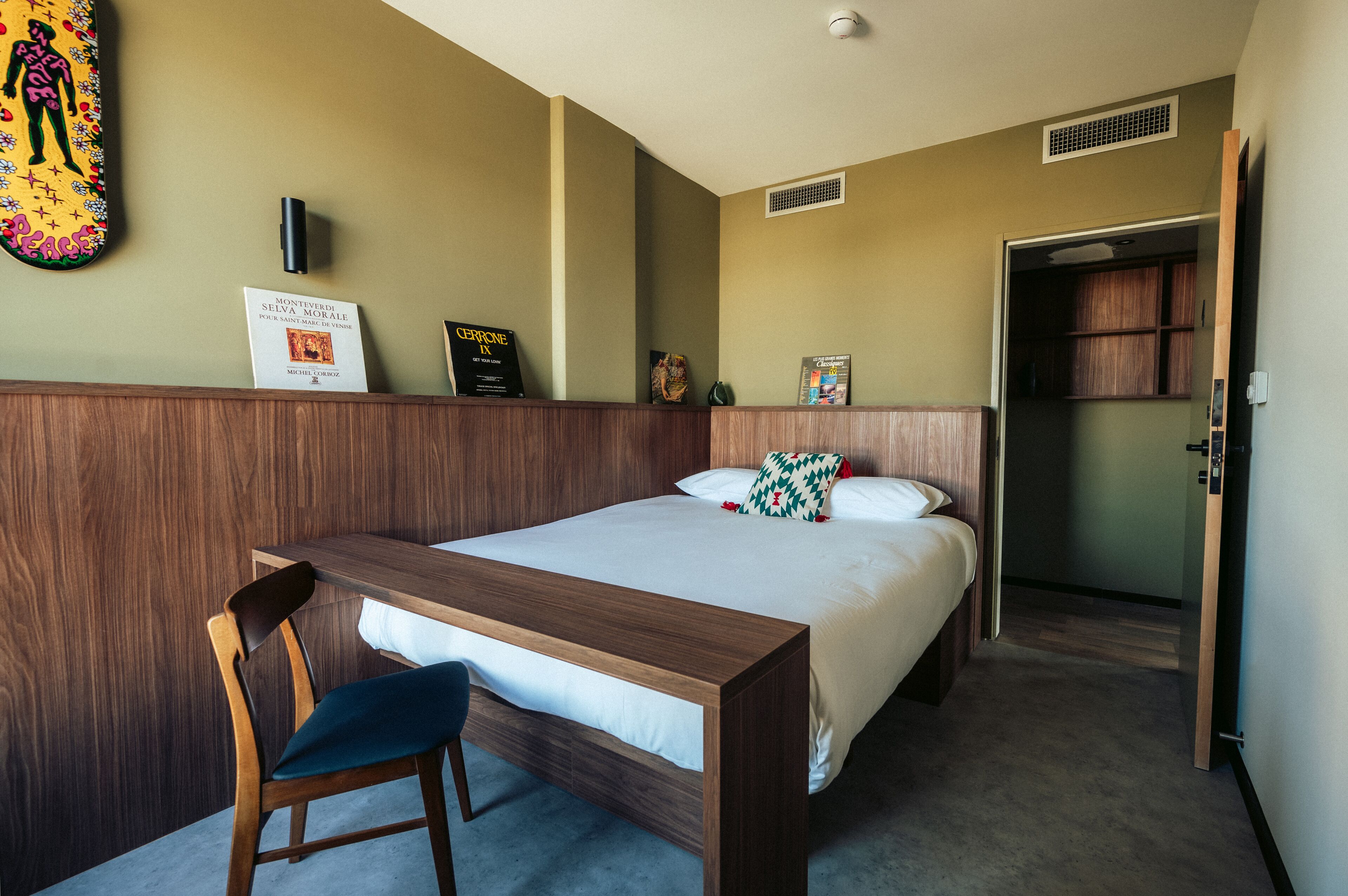 A hotel room with a wooden bed, a desk, and a chair. There is a poster on the wall and a door leading to another room.