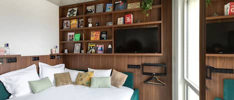 Une chambre d’hôtel avec un grand lit, un téléviseur fixé au mur, une bibliothèque contenant divers objets et une fenêtre donnant sur un beau paysage.