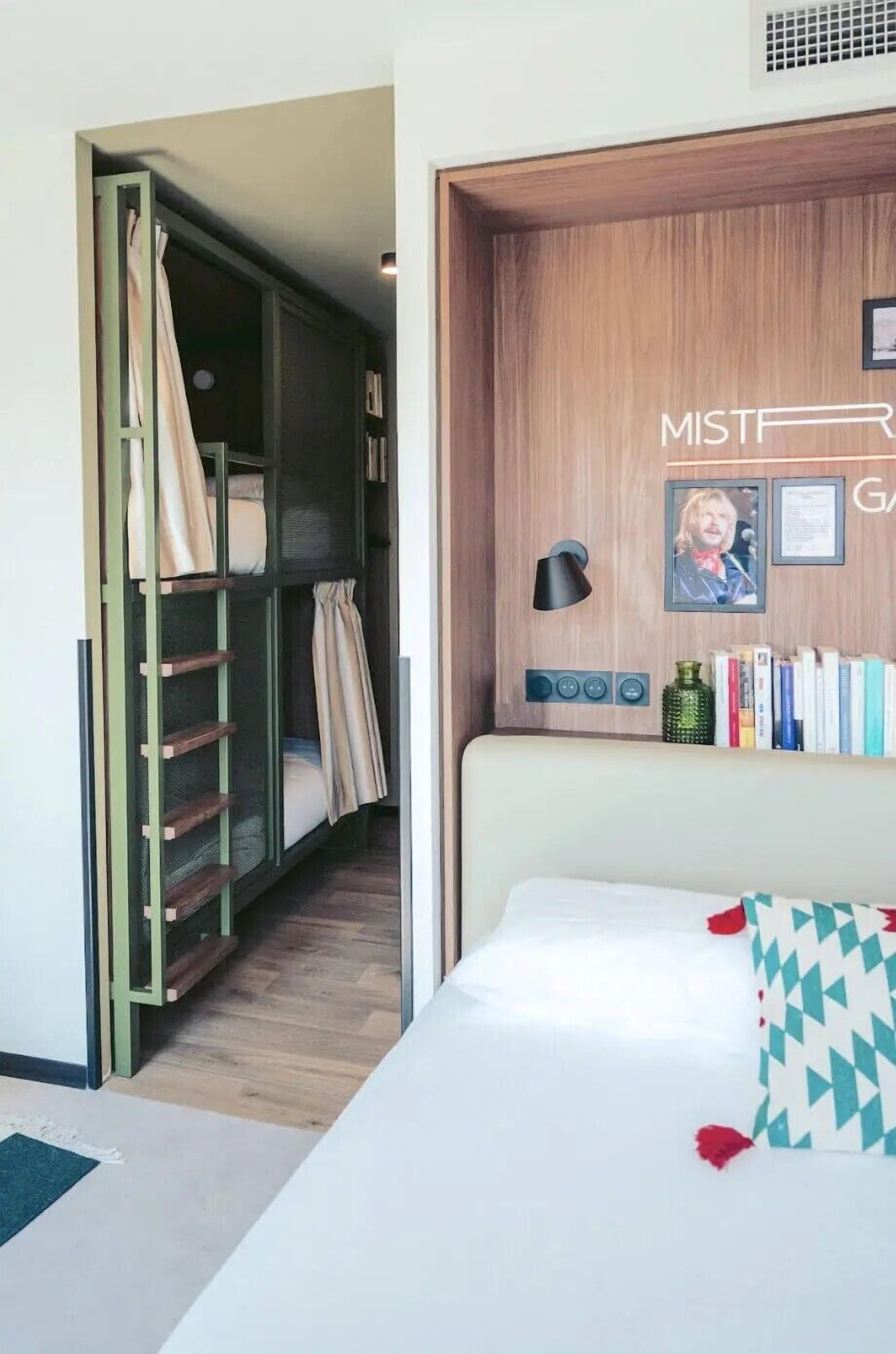 A bedroom with a bunk bed, a bedside table with a lamp, a wall-mounted shelf with books, and a framed picture.