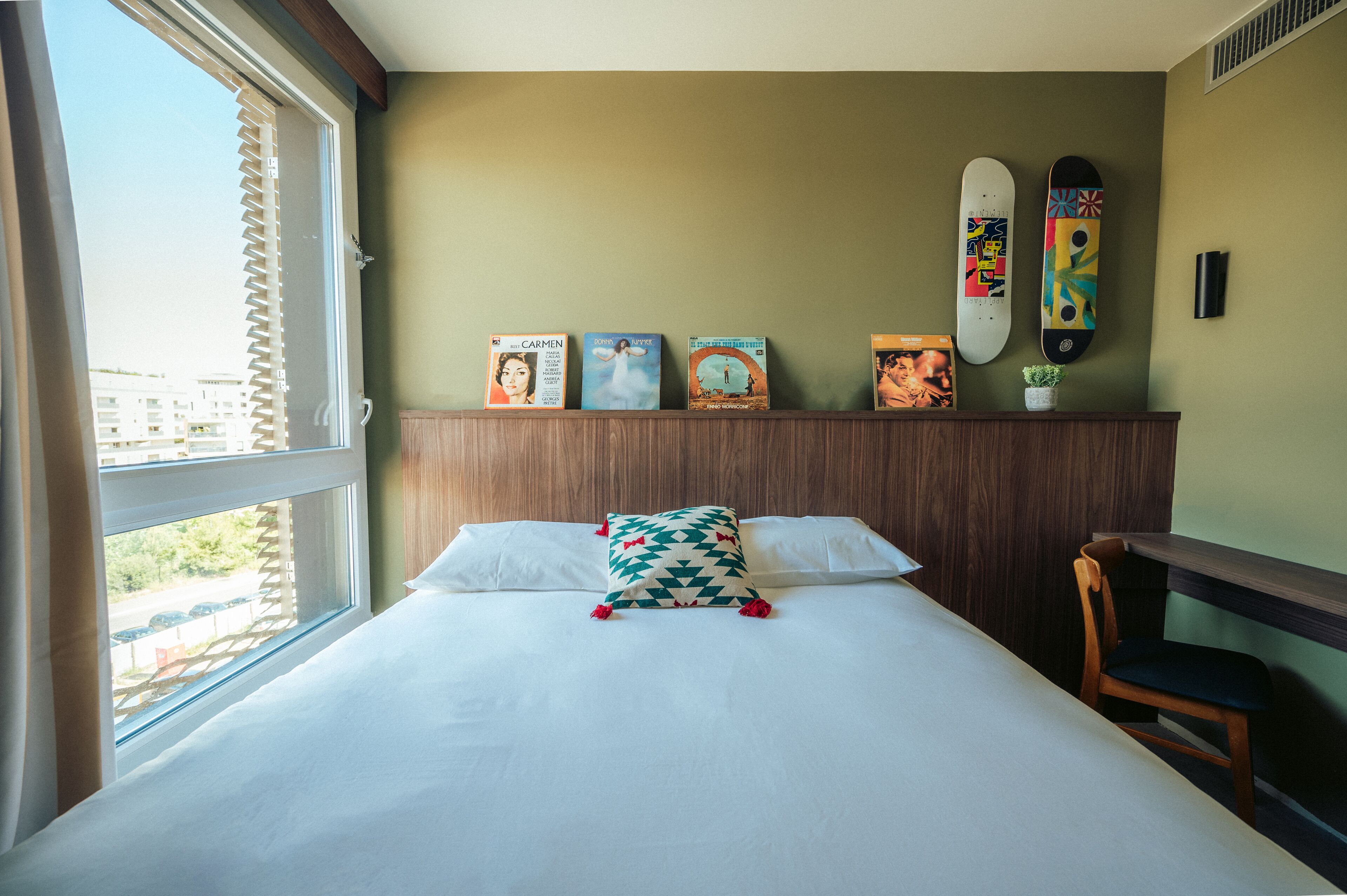 A neatly made bed with a pillow, a wooden headboard, a shelf with framed pictures, and a window with blinds.