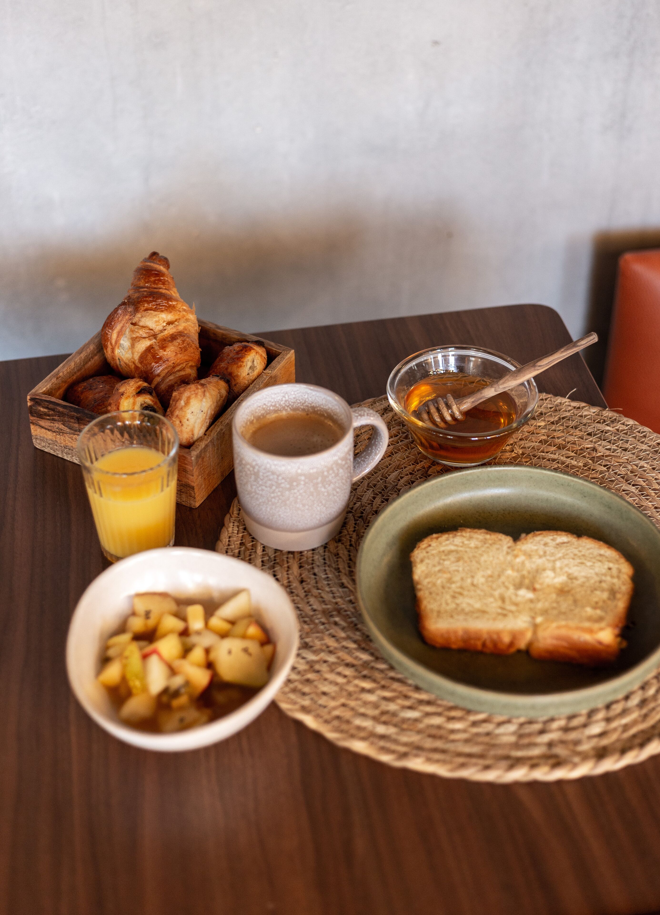 A breakfast table with croissants, toast, a bowl of fruit, a glass of orange juice, a cup of coffee, and a bowl of honey.