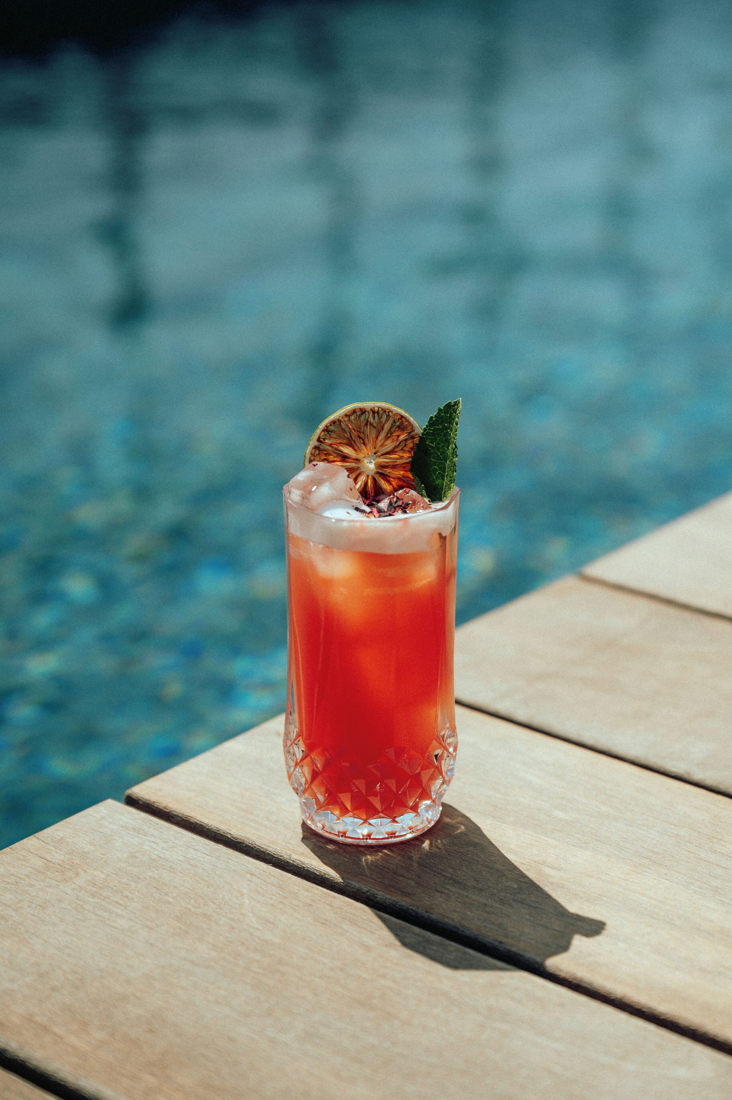 A glass of red beverage with a slice of orange and mint on a wooden table by a pool.