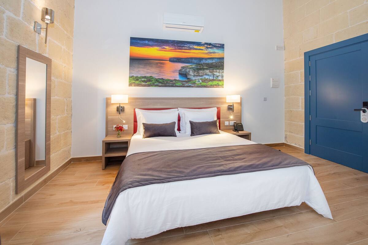 Habitación de hotel con una cama grande, un cuadro de una puesta de sol sobre acantilados, una puerta azul y una pared de piedra.
