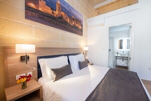 Una cama bien hecha con ropa de cama blanca y gris, una mesita de noche con un florero y un cuadro de un edificio histórico en la pared.