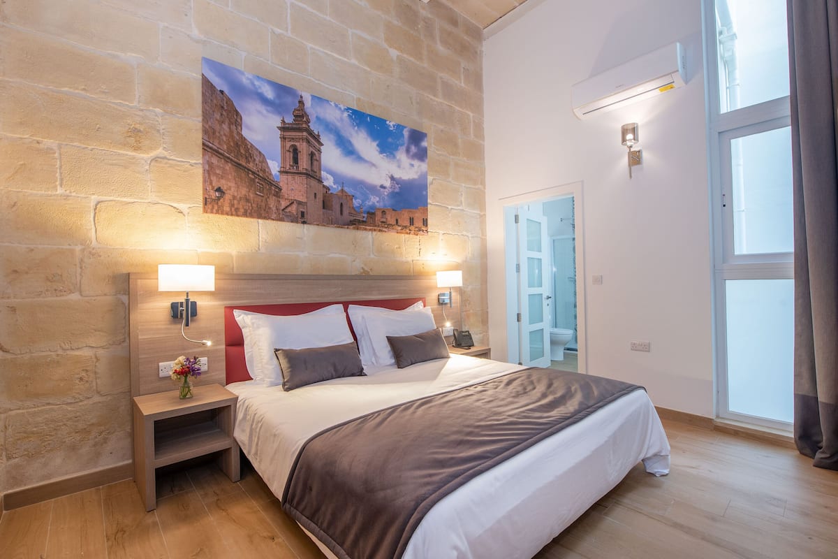 Habitación de hotel con una cama grande, pared de piedra, un cuadro de un edificio histórico, una mesita de noche con una lámpara y una ventana con cortinas.