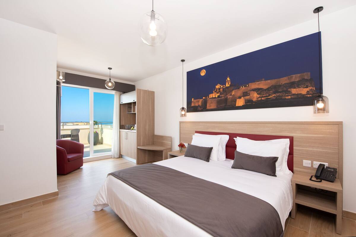 Una habitación de hotel moderna con una cama grande, un sofá rojo, un balcón con vistas y un cuadro de un paisaje urbano.