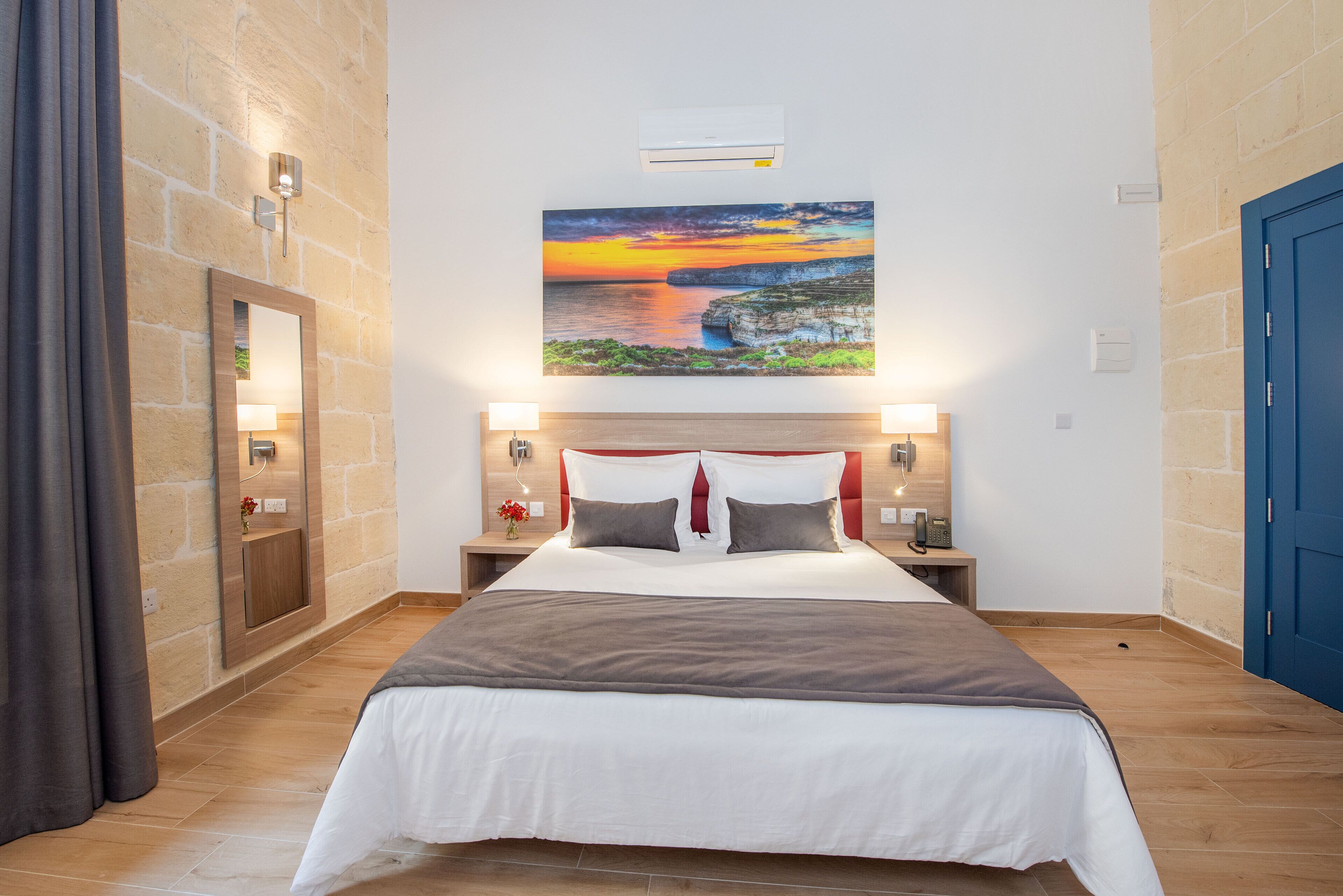 A hotel room with a large bed, a nightstand with a lamp, a wall-mounted air conditioner, and a painting of a sunset over cliffs.