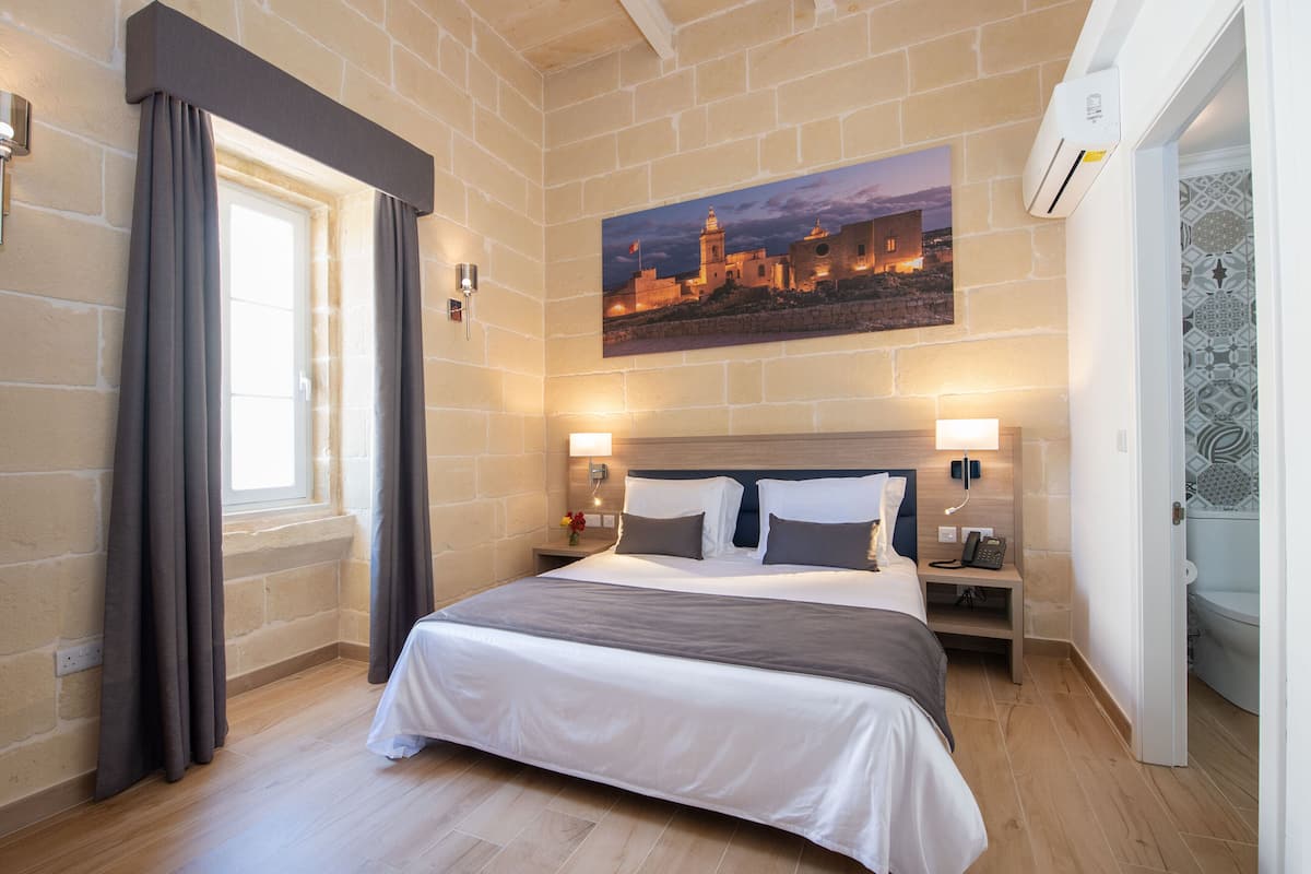 Habitación de hotel con una cama grande, una mesita de noche, una lámpara de pared, un cuadro de un paisaje urbano y una puerta que da al baño.