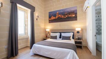 A hotel room with a large bed, a nightstand, a wall-mounted lamp, a painting of a cityscape, and a door leading to the bathroom.