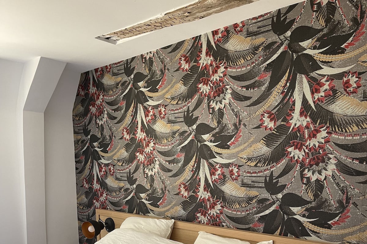 A bedroom with a large bed, a headboard with a floral pattern, a bedside table with a lamp, and a small shelf.