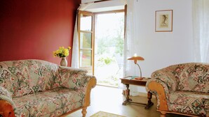 A room with two floral-patterned sofas, a wooden table with a lamp, and a window with curtains. - LE BOS CLARD farm large garden, terras, 9pp, (Felines)