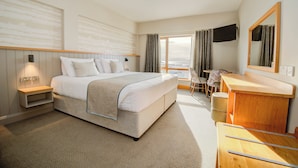 A hotel room with a bed, bedside table, desk, chair, and a large window with curtains. - Beach Hotel (Downings)