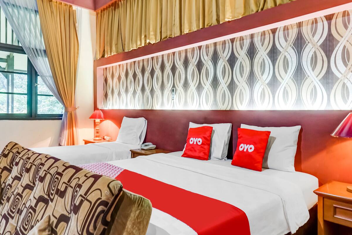 A hotel room with two beds, a patterned throw blanket, a decorative wall, and a window with curtains.