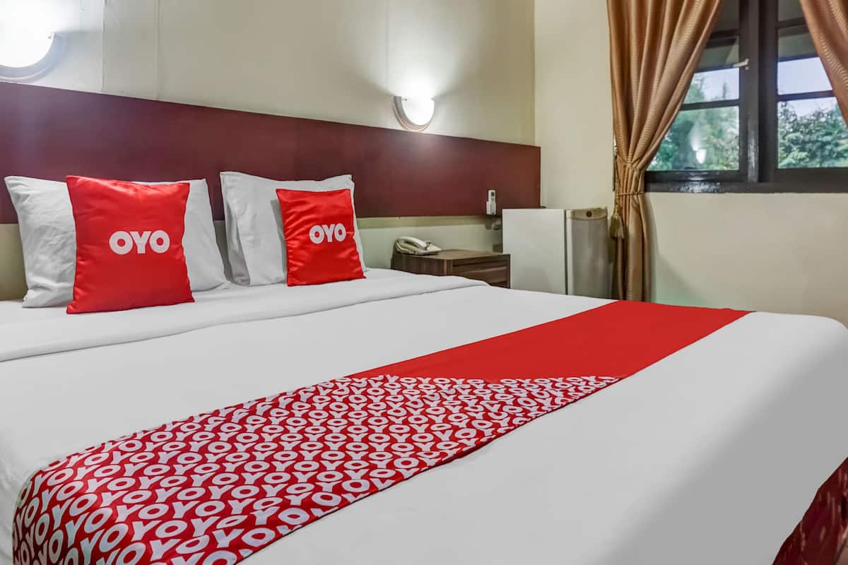 A hotel room with a bed, two red pillows with 'OYO' branding, a red and white patterned bedspread, a nightstand with a phone, and a window with curtains.
