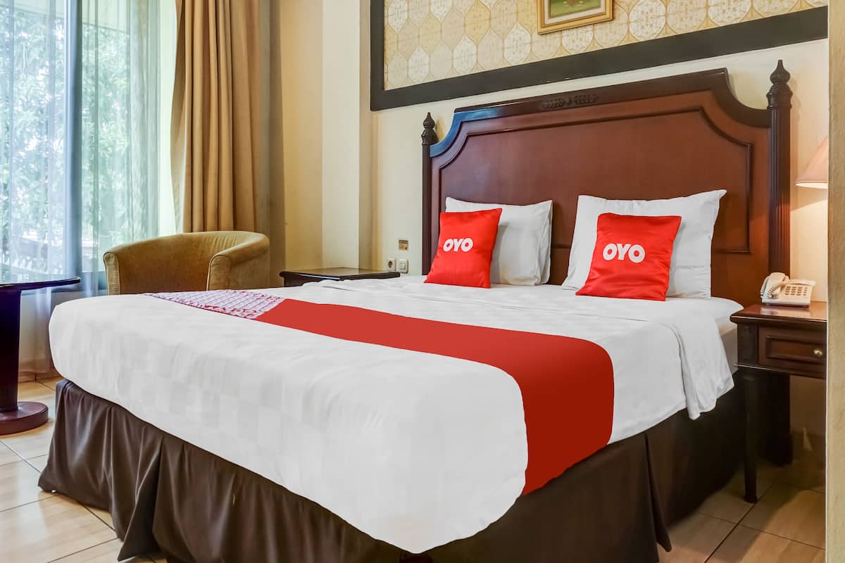 A hotel room with a large bed, two red pillows with "OYO" branding, a wooden headboard, a bedside table with a lamp, and a view of greenery through the window.