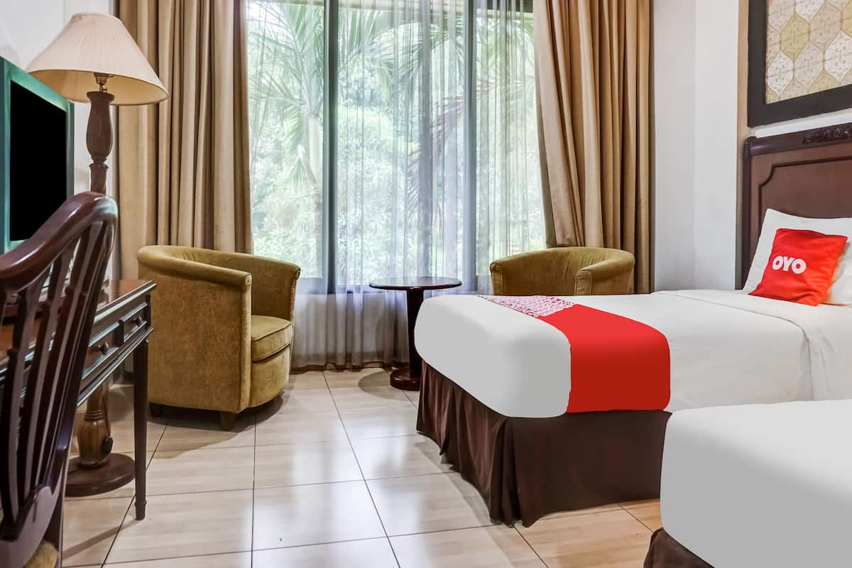 A hotel room with two single beds, a desk with a lamp, two armchairs, a small table, and a large window with curtains.