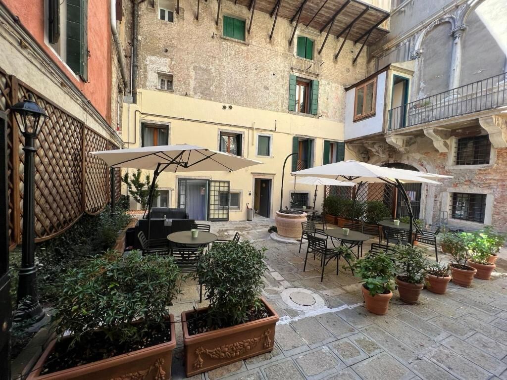Un patio exterior con mesas y sillas, rodeado de plantas en macetas y un suelo de piedra.