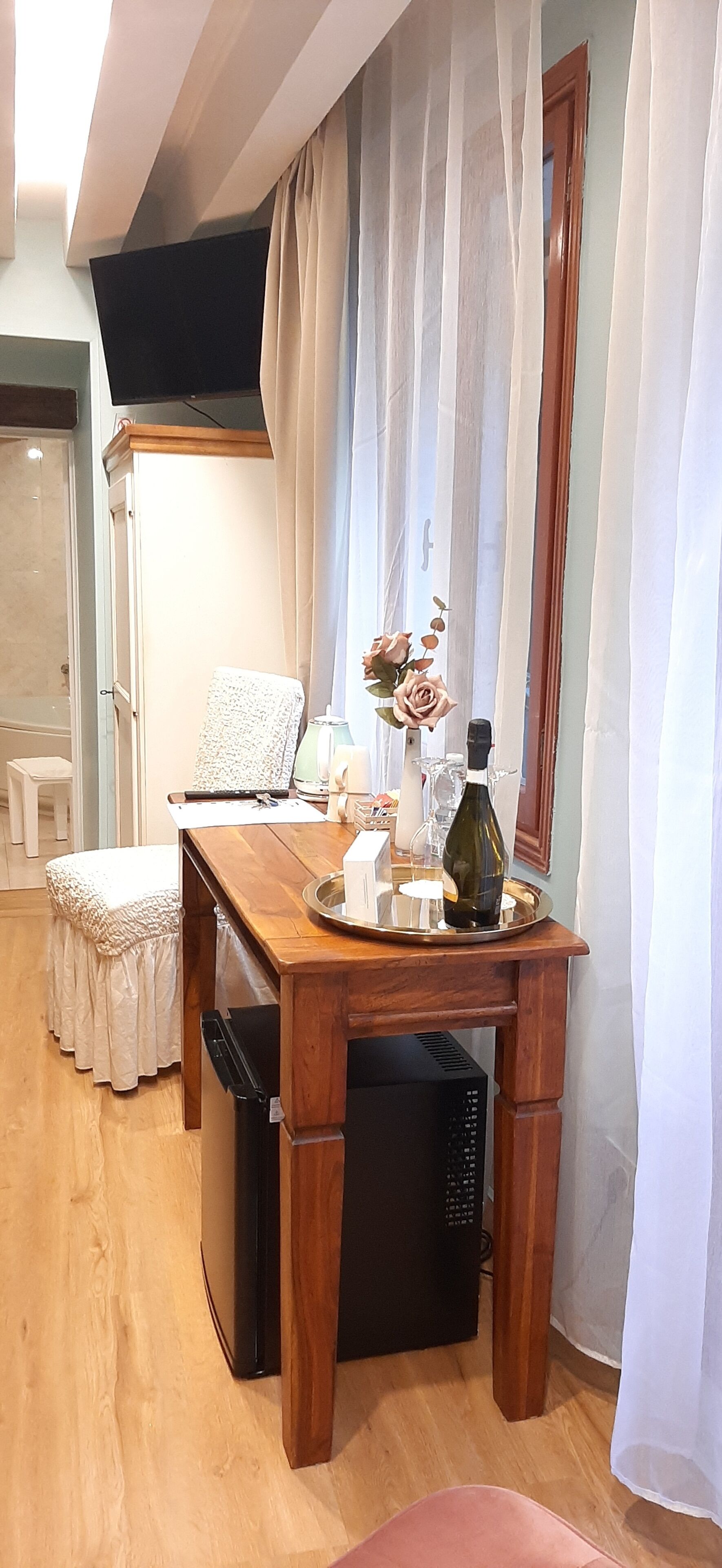 Een houten eettafel met een dienblad, een fles en een vaas met bloemen. Er staat een witte stoel en een klein wit tafeltje met een handdoek. Een zwart apparaat staat onder de tafel.