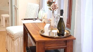 Una mesa de madera para comer con una bandeja, una botella y un florero con flores. Hay una silla blanca y una mesita pequeña también blanca, con una toalla. Un electrodoméstico negro está debajo de la mesa.
