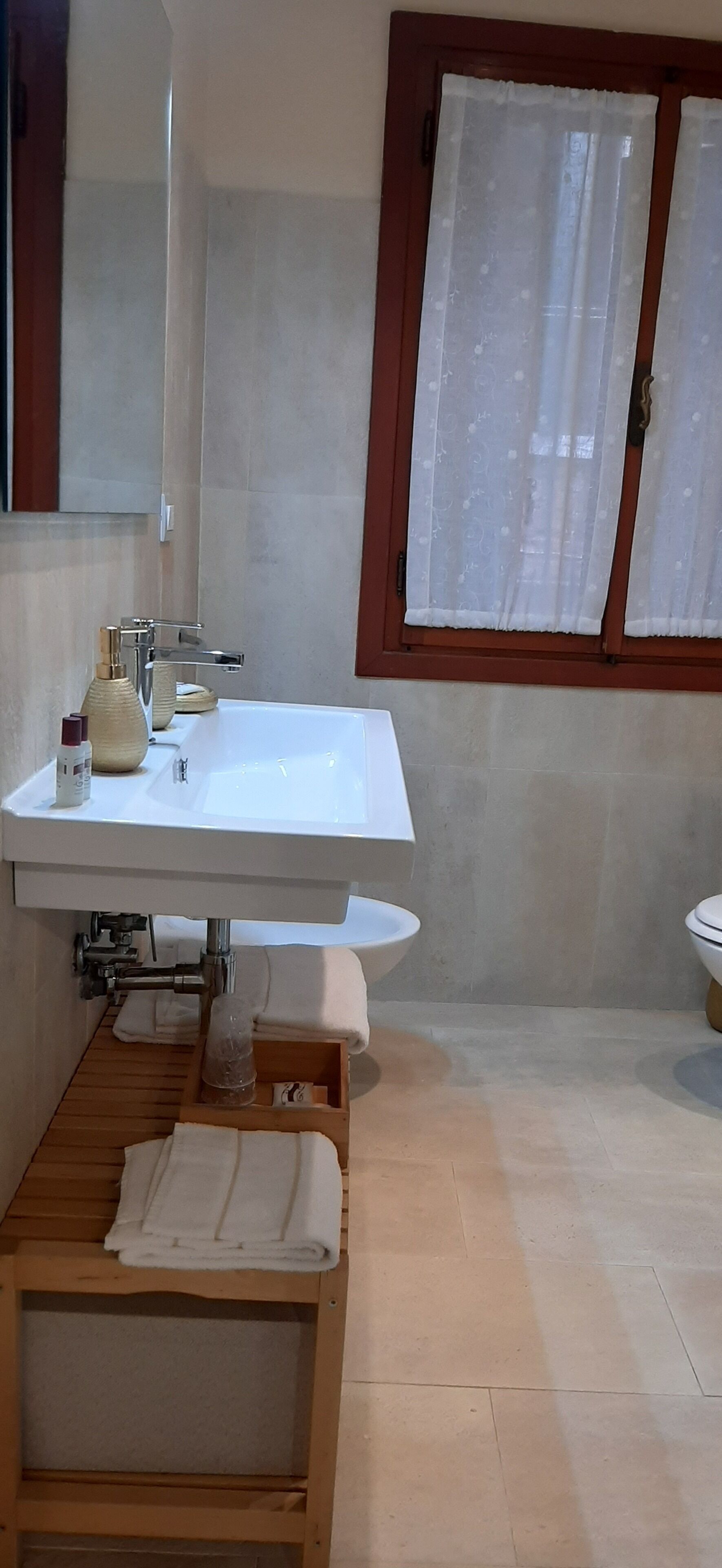 Un baño con lavamanos blanco, un espejo, una ventana con cortina blanca y un estante de madera con toallas.