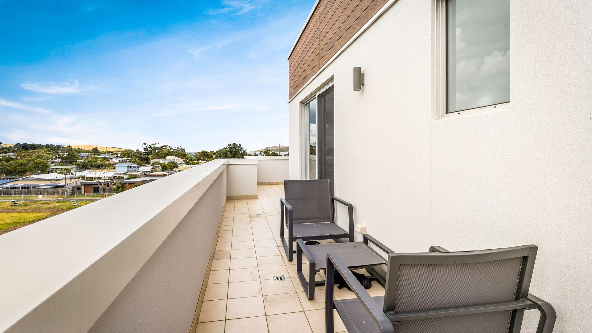 A balcony with two chairs and a table, a white wall, and a view of houses and hills in the background.