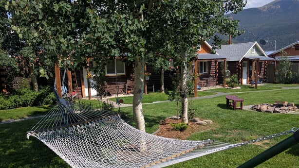 Un hamac dans un espace verdoyant, avec une terrasse en bois et un chalet en arrière-plan.