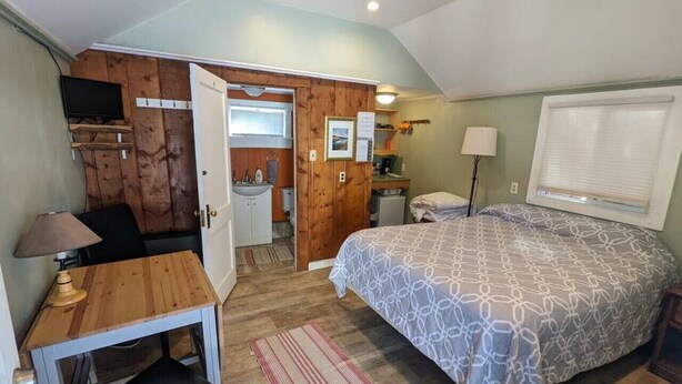 Quarto com uma cama, escrivaninha, cadeira, abajur, janela com persianas e porta que dá para um banheiro.