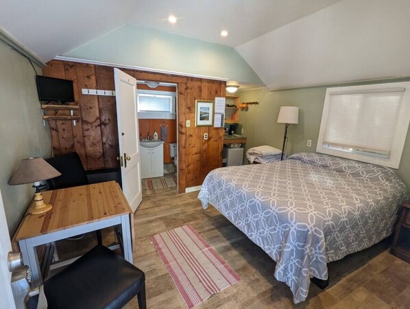 A bedroom with a bed, a desk, a chair, a lamp, a window with blinds, and a door leading to a bathroom.