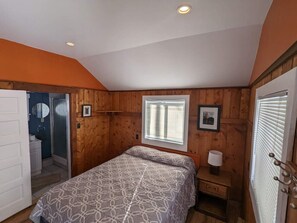 Een slaapkamer met een bed, een raam, een nachtkastje met een lamp en een deur naar een badkamer.