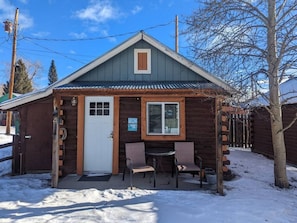 A wooden cabin with a door, two windows, and a small porch with chairs.
