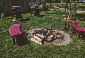 A backyard with a fire pit surrounded by logs, two red Adirondack chairs, and a barbecue grill.