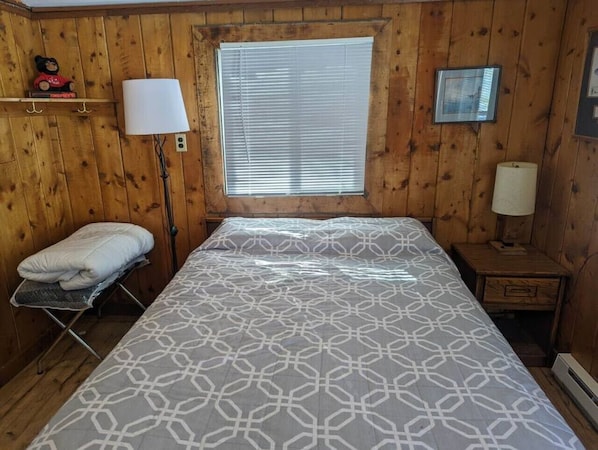 A wooden-paneled room with a bed, a bedside lamp, a window with blinds, and a small shelf with a teddy bear.
