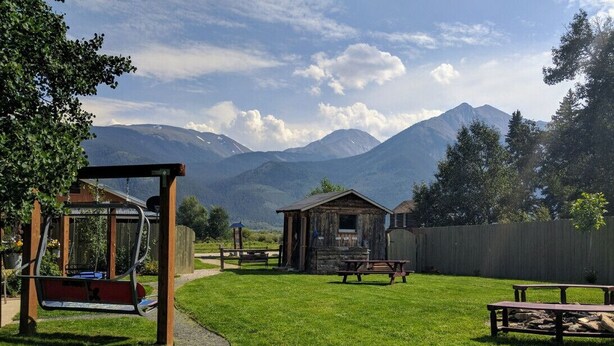 A rustic outdoor setting with a swing set, picnic tables, and a small cabin, surrounded by a wooden fence and a mountain backdrop. - Stay Twin Lakes Cabin 3 (Twin Lakes)