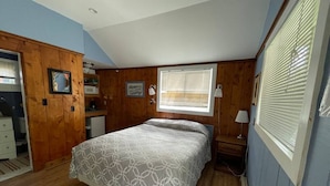 A bedroom with a bed, wooden walls, a window with blinds, a nightstand, and a desk with a lamp. - Stay Twin Lakes Cabin 3 (Twin Lakes)