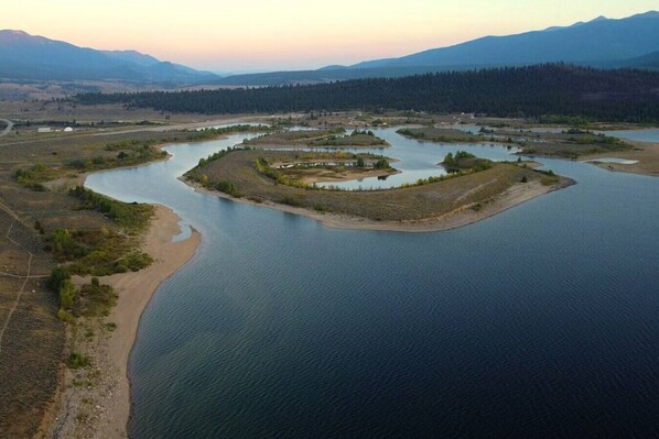 An aerial view of a lake with a small island featuring a circular structure, surrounded by forested hills and mountains.