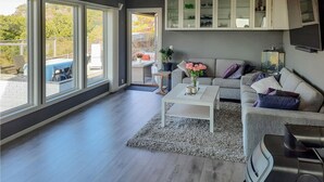 Ett modernt vardagsrum med en grå soffa, ett vitt soffbord och ett stort fönster med utsikt över en trädgård.