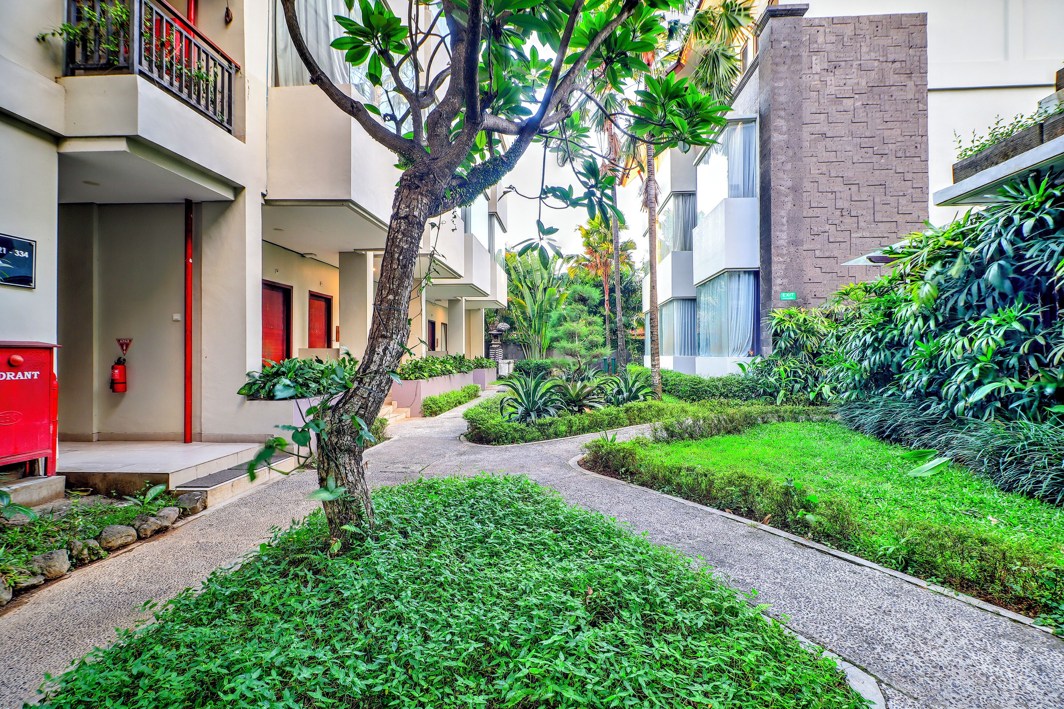 A courtyard with a pathway, greenery, and buildings with balconies and doors.