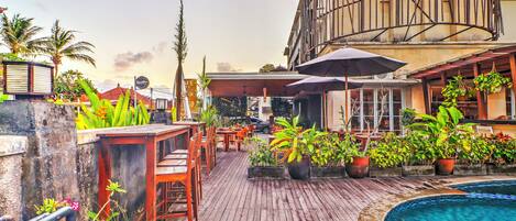 A rooftop bar with wooden flooring, potted plants, outdoor seating, and a pool.