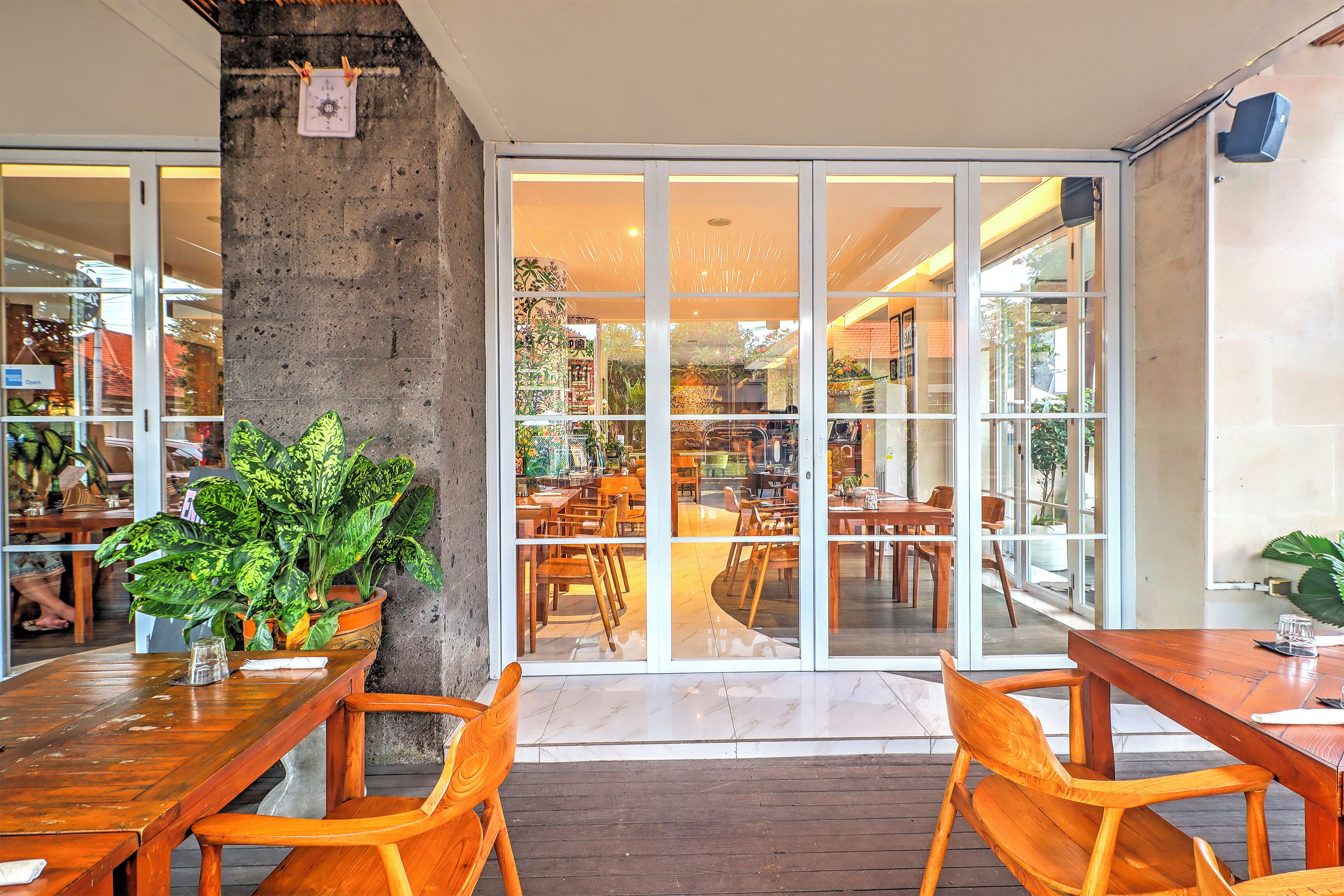 A restaurant with wooden tables and chairs, a potted plant, and a view through glass doors into the dining area.